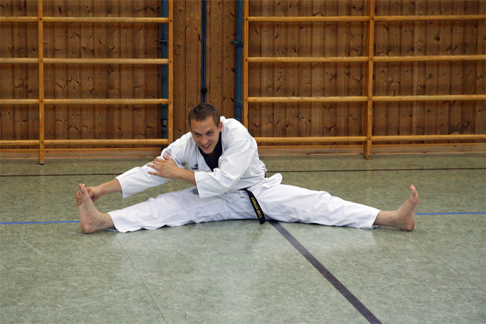 Spagat-Dehnung von einem sitzenden Taekwondo-Schüler mit
        gespreitzten Beinen, der mit der Hand seinen rechten Fuß
        umfasst